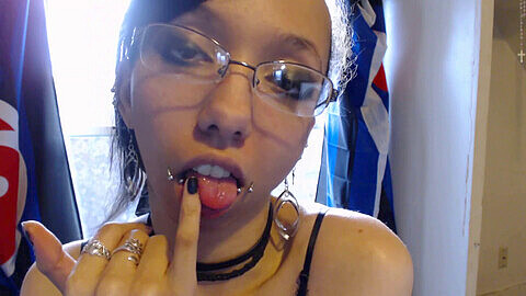 Ein Emo-Babe mit Piercings neckt einen Glasdildo mit ihrer Zunge, nachdem sie rosa Lipgloss aufgetragen hat.
