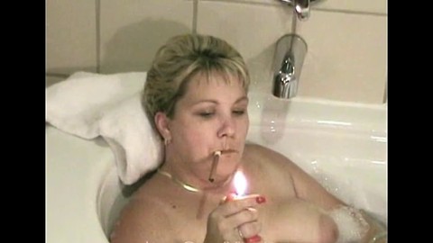 Une milf chaude prend un bain et fume un cigare avec de larges aréoles