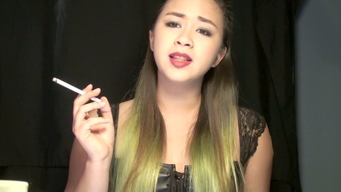 Fumeuse asiatique alternative se livrant à sa passion en portant un patch de nicotine 21mg et en fumant deux cigarettes tout en dominant son esclave soumis habillé d'un collier.